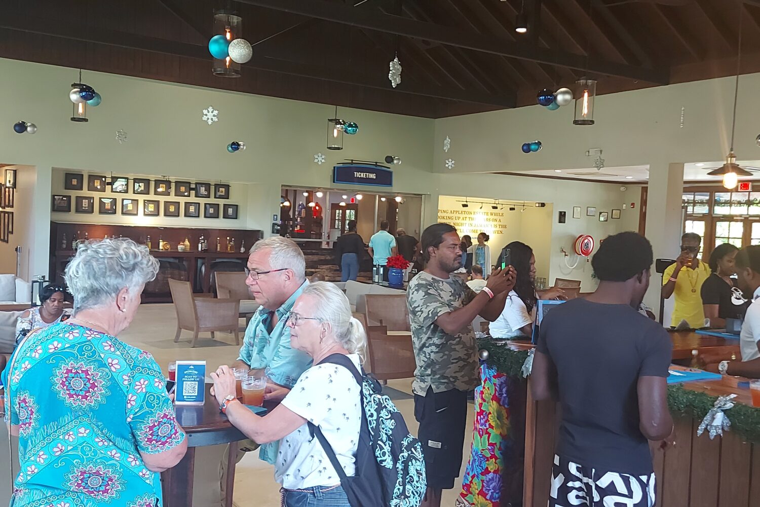 Groups of visitors sit at tables and around the bar in the Appleton Estate reception area, sipping rum-based beverages and socializing while waiting for their tour guide to begin the factory tour.