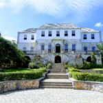 Rose Hall Great House in Montego Bay, Jamaica, viewed from the paved walkway with steps leading up to the historic Georgian mansion, surrounded by manicured lawns and tropical trees.