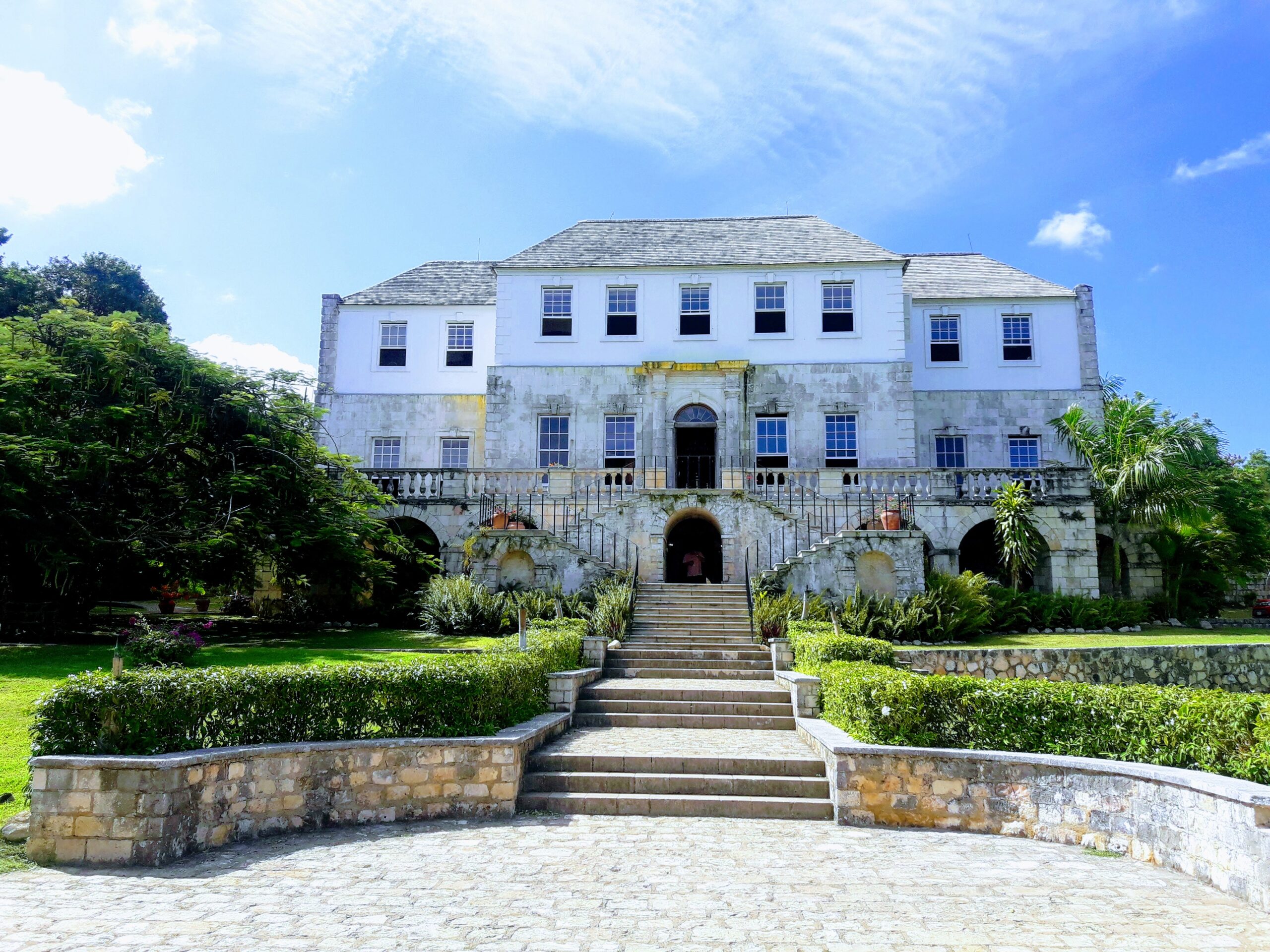 Rose Hall Great House in Montego Bay, Jamaica, viewed from the paved walkway with steps leading up to the historic Georgian mansion, surrounded by manicured lawns and tropical trees.