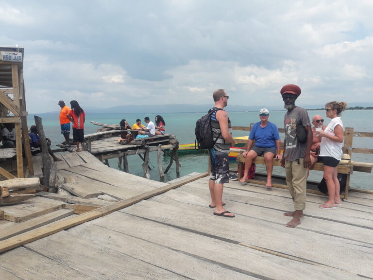 People standing and conversing, sitting on a wooden bench, and looking into the water on the deck of the Pelican Bar, surrounded by the Caribbean Sea.
