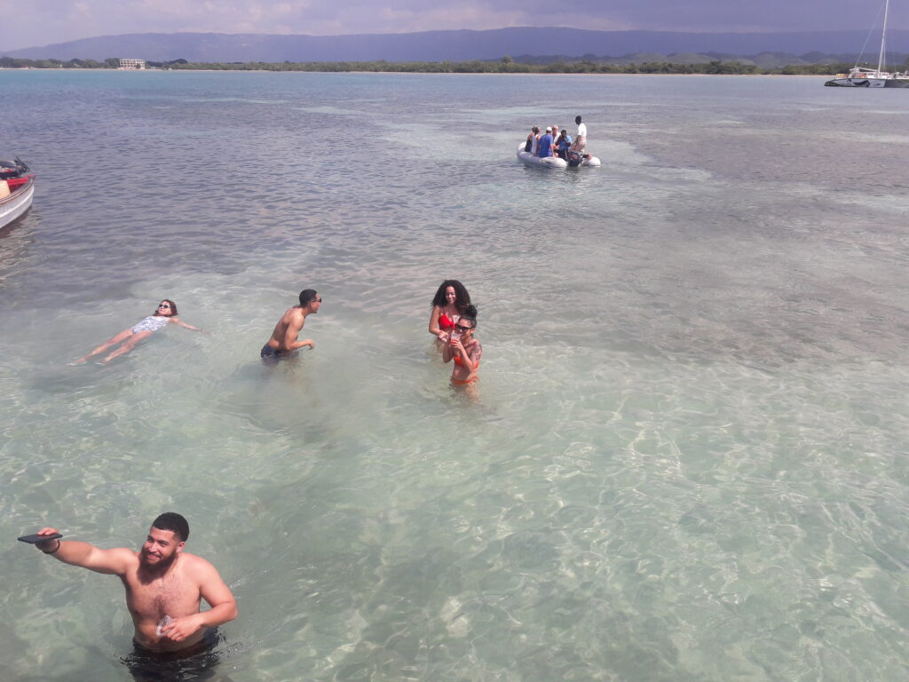 Visitors swimming in the shallow Caribbean waters at Pelican Bar, sipping Red Stripe beer and other beverages, enjoying Jamaica’s unique sandbar bar experience.