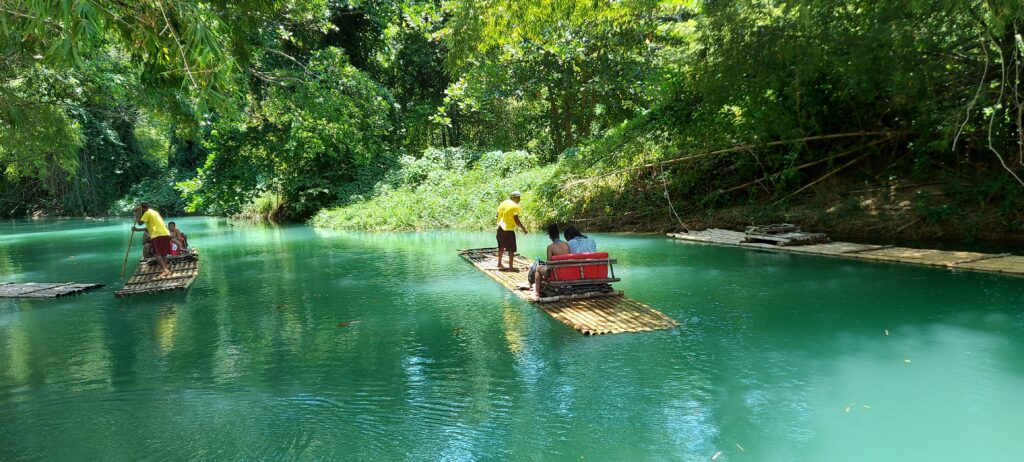 2 people seated and1 man standing on a bamboo raft floating on a river