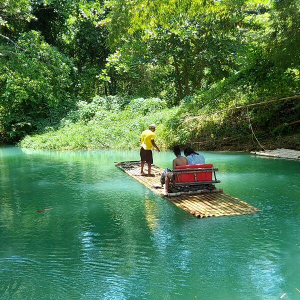 2 people seated and1 man standing on a bamboo raft floating on a river