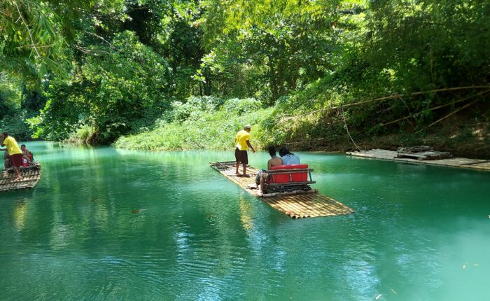 2 people seated and1 man standing on a bamboo raft floating on a river