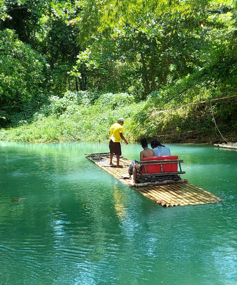 2 people seated and1 man standing on a bamboo raft floating on a river