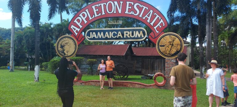 A couple poses at the Appleton Estate picture stand as the tour guide takes their photo, while other visitors watch nearby.