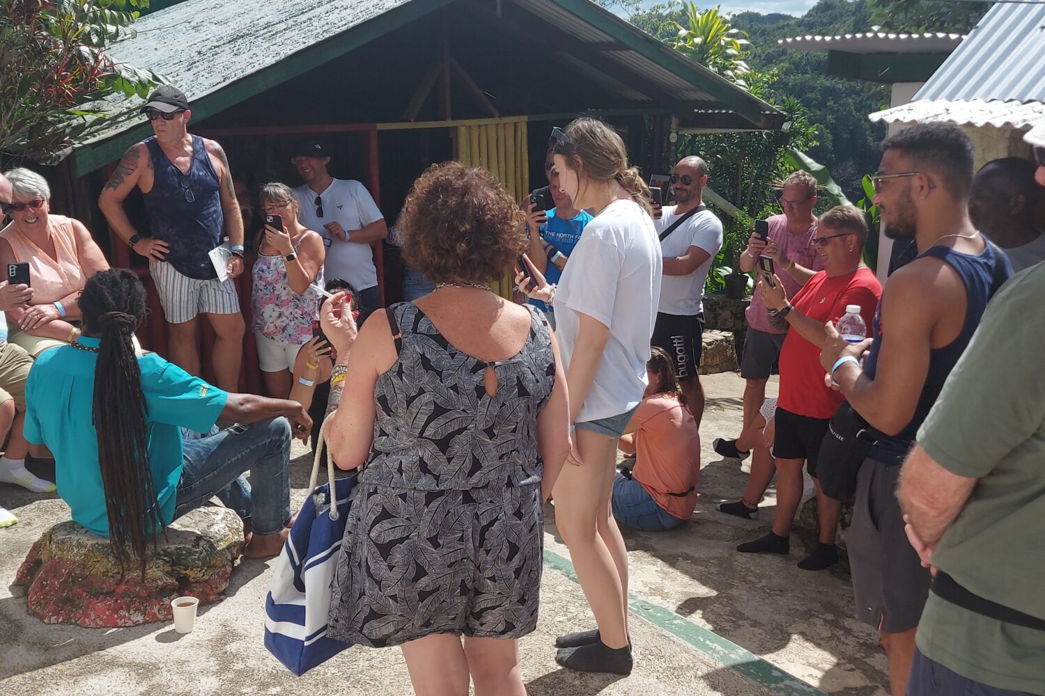 A group of visitors surround a tour guide seated on the ground in the yard at Nine Mile, listening to stories about Bob Marley