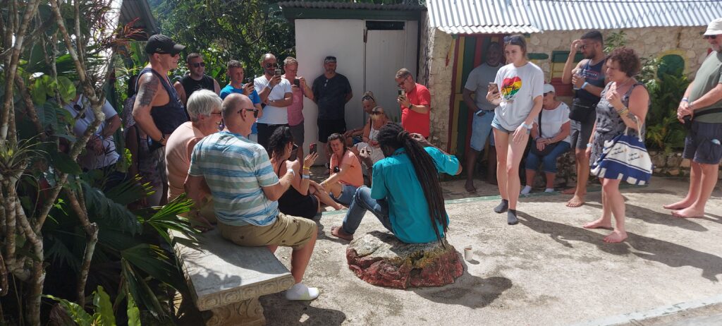 Tour guide with locks sits on a stone in the yard at Nine Mile, as visitors sit and stand around him listening to information about Bob Marley.