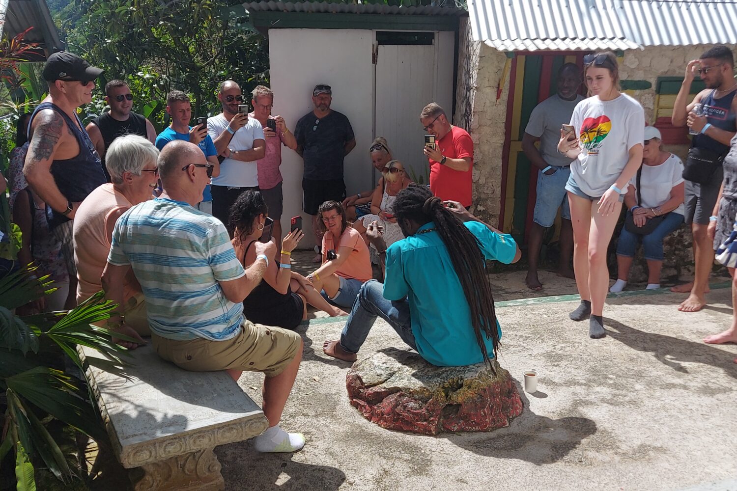 Tour guide with locks sits on a stone in the yard at Nine Mile, as visitors sit and stand around him listening to information about Bob Marley.