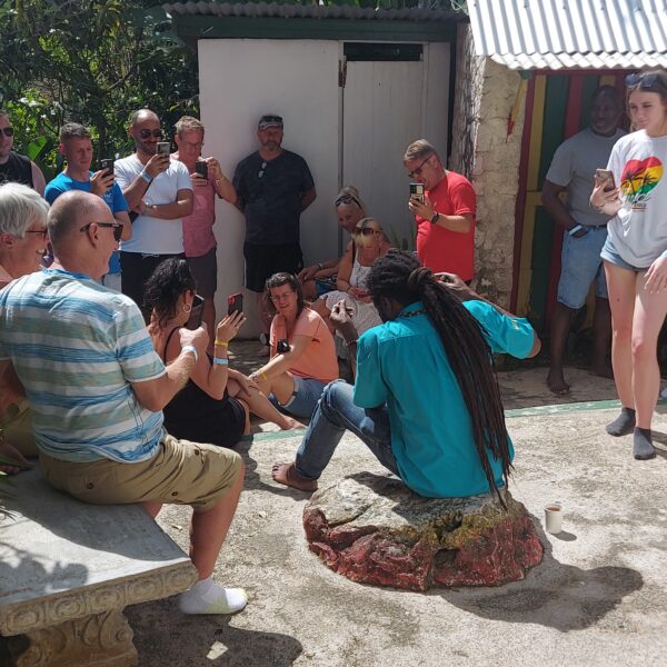 Tour guide with locks sits on a stone in the yard at Nine Mile, as visitors sit and stand around him listening to information about Bob Marley.