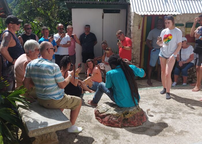 Tour guide with locks sits on a stone in the yard at Nine Mile, as visitors sit and stand around him listening to information about Bob Marley.
