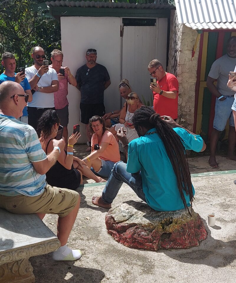 Tour guide with locks sits on a stone in the yard at Nine Mile, as visitors sit and stand around him listening to information about Bob Marley.