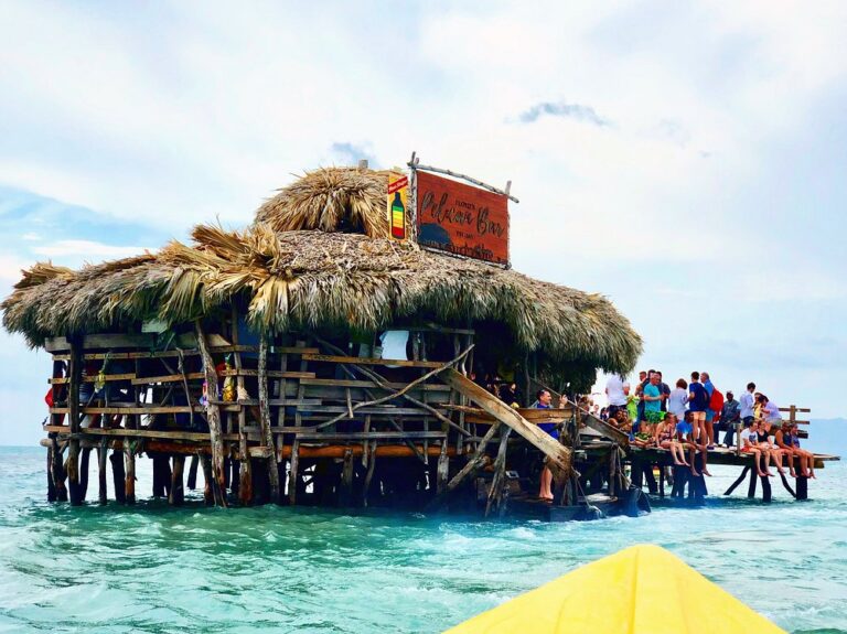 Front view of the Pelican Bar with its sign on top and a group of visitors sitting on the wooden deck above the water