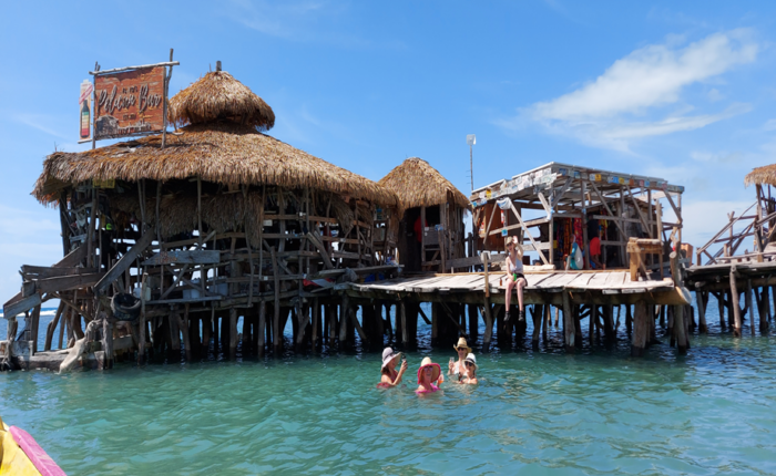 People relaxing on the wooden deck of the Pelican Bar while others swim in the shallow turquoise water surrounding the rustic driftwood structure