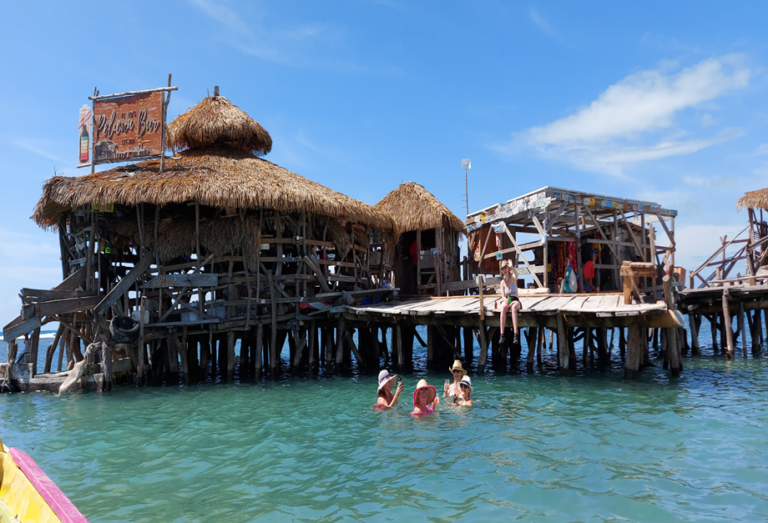 People relaxing on the wooden deck of the Pelican Bar while others swim in the shallow turquoise water surrounding the rustic driftwood structure