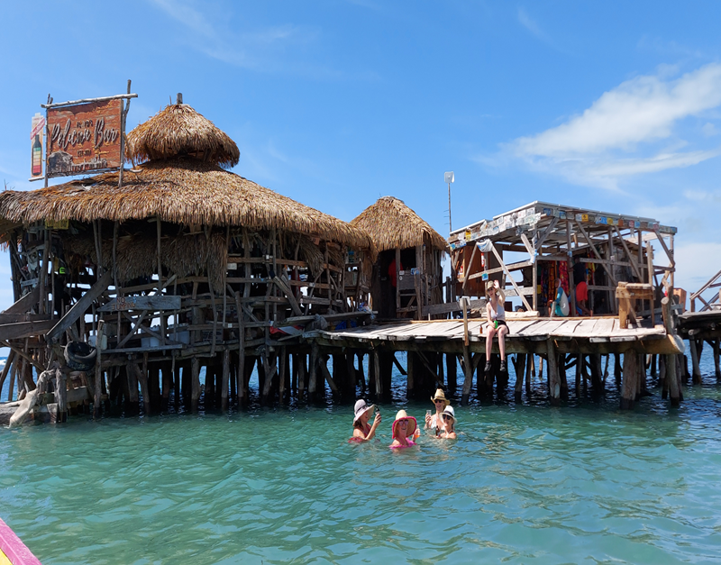 People relaxing on the wooden deck of the Pelican Bar while others swim in the shallow turquoise water surrounding the rustic driftwood structure