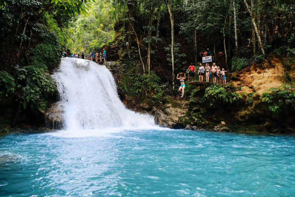 Visitors and a guide stand atop Blue Hole Falls while others swim and watch from rocks below.