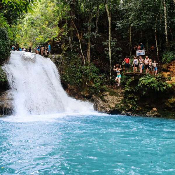 Visitors and a guide stand atop Blue Hole Falls while others swim and watch from rocks below.