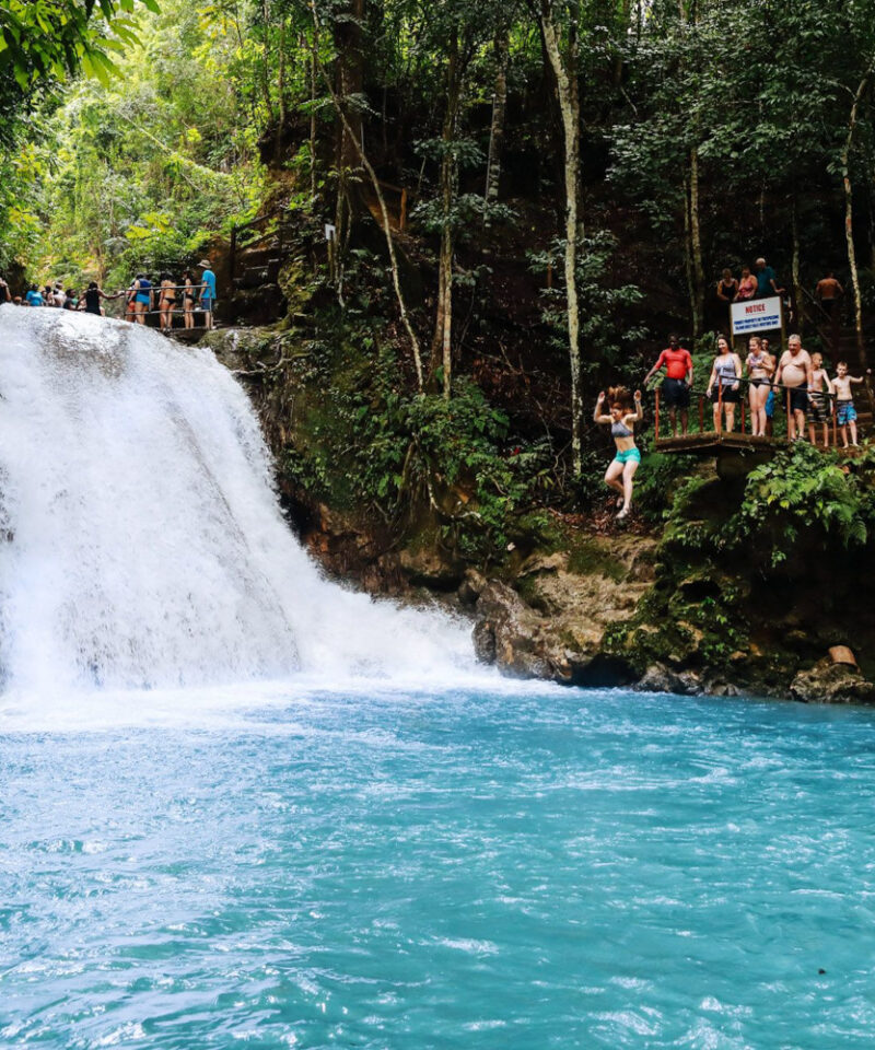 Visitors and a guide stand atop Blue Hole Falls while others swim and watch from rocks below.