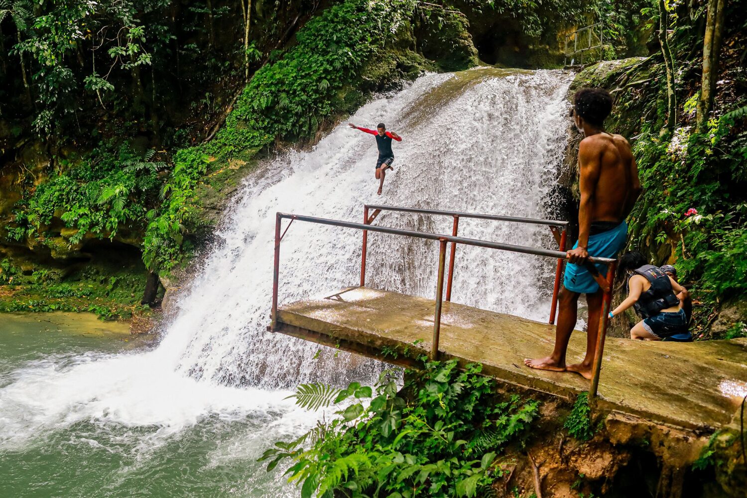 Tour guide walks halfway down Blue Hole Falls while another guide looks on; visitors swim below.