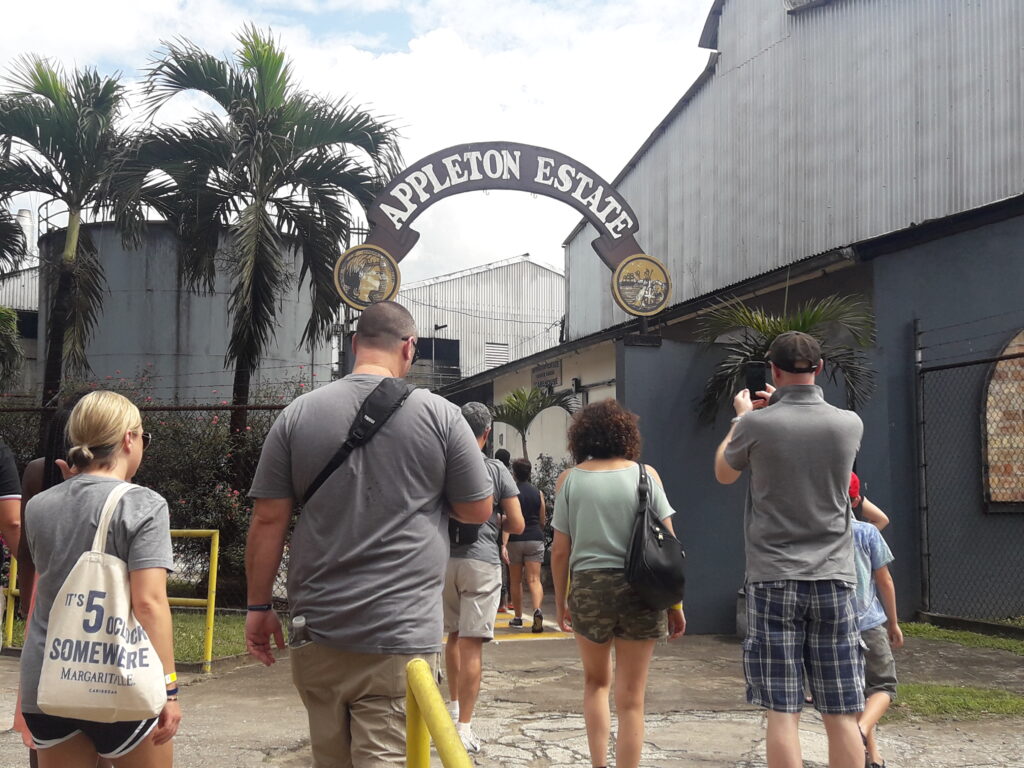 Visitors walk toward the entrance gate of Appleton Estate, where an arch overhead displays the Appleton Estate sign. Some guests pause to take photos as they approach, while others continue toward the factory for the guided tour.