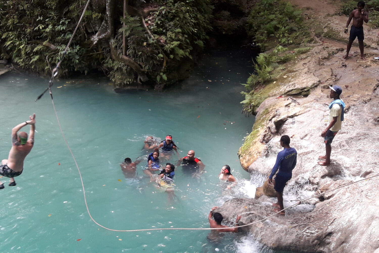 A group of visitors swim in the turquoise waters of the Blue Hole, looking up as another visitor swings on a rope from the rocks above, preparing to plunge into the pool.