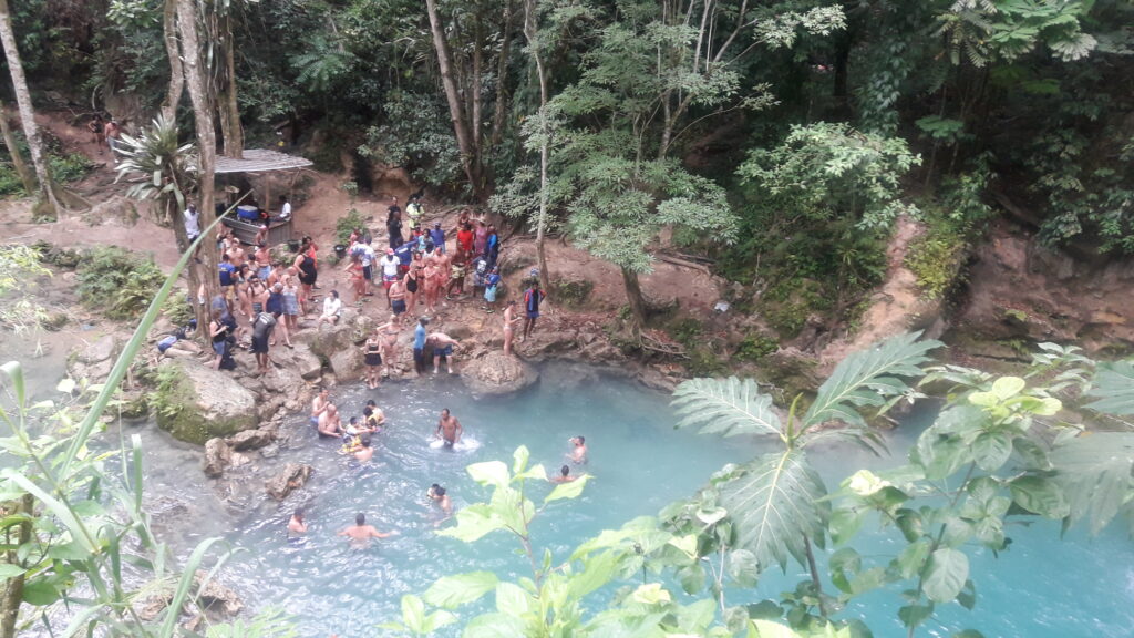 Visitors swim in Blue Hole river while others rest on rocks among lush greenery.