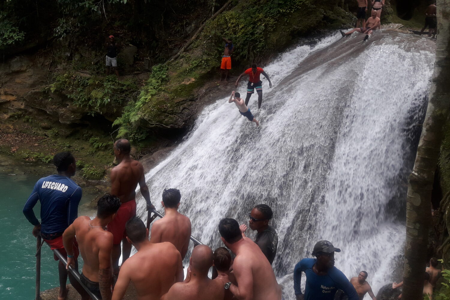 Visitors stand on rocks across from the cascading Blue Hole falls, watching as a guide assists another visitor preparing to jump into the turquoise pool below.
