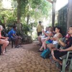 Visitors sit under a wooden gazebo at Rocklands Bird Sanctuary, holding small bottles of nectar as they wait for hummingbirds to perch on their fingers and drink. A tour guide sits nearby, ready to take photos and share insights, creating an unforgettable travel experience.