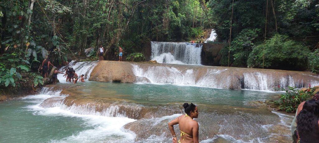 Seven-tiered YS Falls cascading over rocky ledges, with water flowing down as one visitor looks on from the top and others walk across a shallow section of the stream below.
