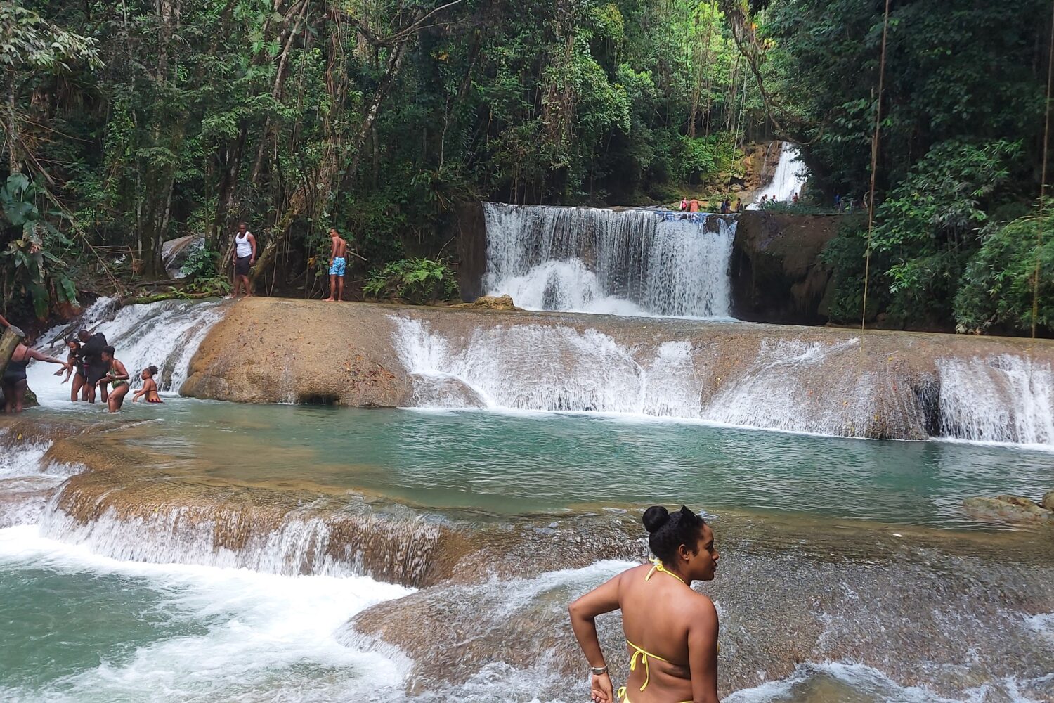 Seven-tiered YS Falls cascading over rocky ledges, with water flowing down as one visitor looks on from the top and others walk across a shallow section of the stream below.