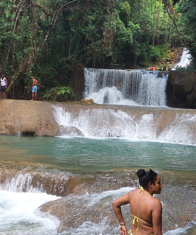 Seven-tiered YS Falls cascading over rocky ledges, with water flowing down as one visitor looks on from the top and others walk across a shallow section of the stream below.