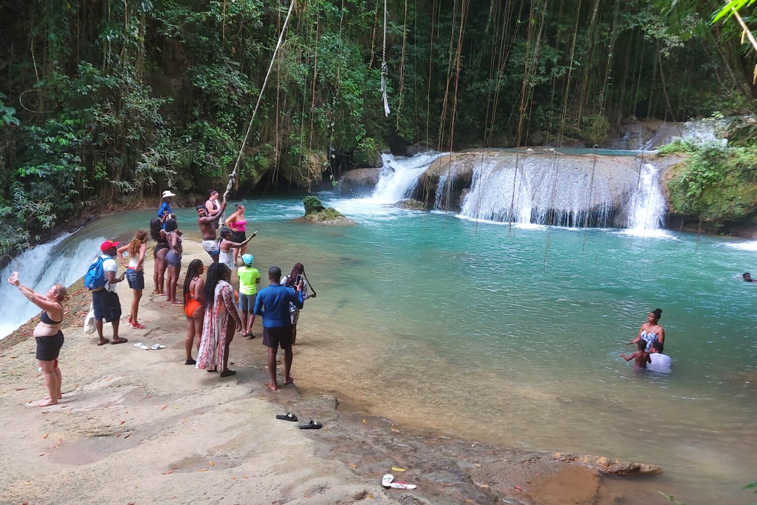 Visitors enjoying YS Falls, with some swimming in the natural pools, others swinging on the rope swing, and several people standing nearby taking photos and watching the cascading waterfall.