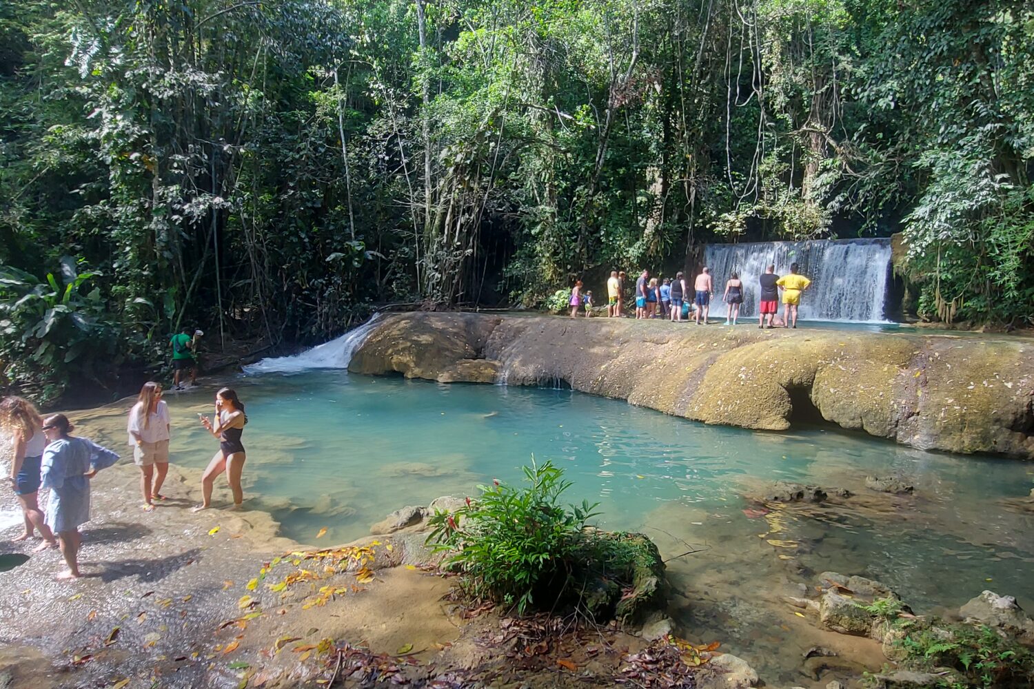 Two visitors stand at the foot of YS Falls before a deep blue pool, taking photos of the cascading waterfall ahead, while other guests walk across different tiers above.