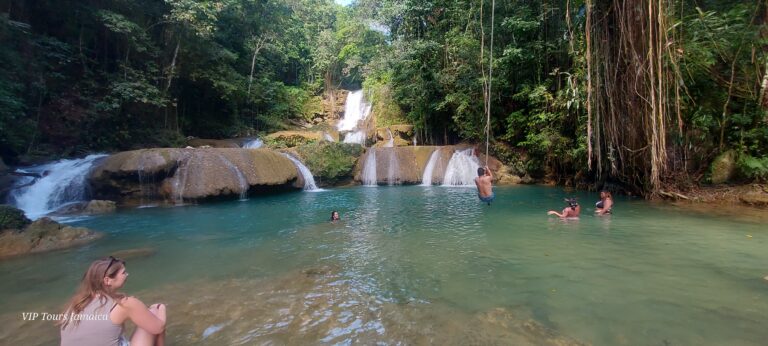 A visitor sits in the water at the first tier of YS Falls, gazing at the full seven-tier waterfall, while other visitors walk across the top tier and some stand nearby observing the scene.