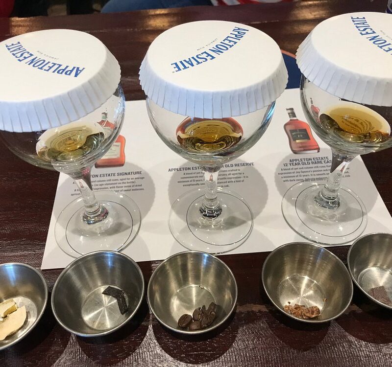Three glasses of rum, each covered with white Appleton-branded disposable lids, sit on a tasting room table at Appleton Estate. Beside them are five stainless steel containers holding different spices, arranged neatly for visitors to sample during the guided tasting experience.