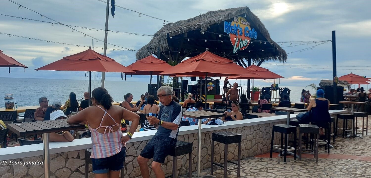 Visitors sit around tables at the cliffside of Rick’s Café in Negril, Jamaica, enjoying live entertainment as the Caribbean Sea provides a stunning backdrop.