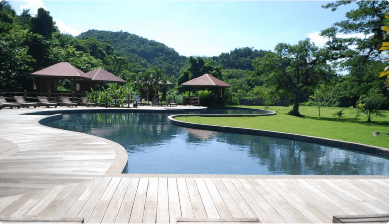 Wide-angle view of the natural river-fed swimming pool at YS Falls, featuring stylish designs and gazebos surrounded by manicured lawns and lush gardens, with majestic mountains rising in the backdrop.