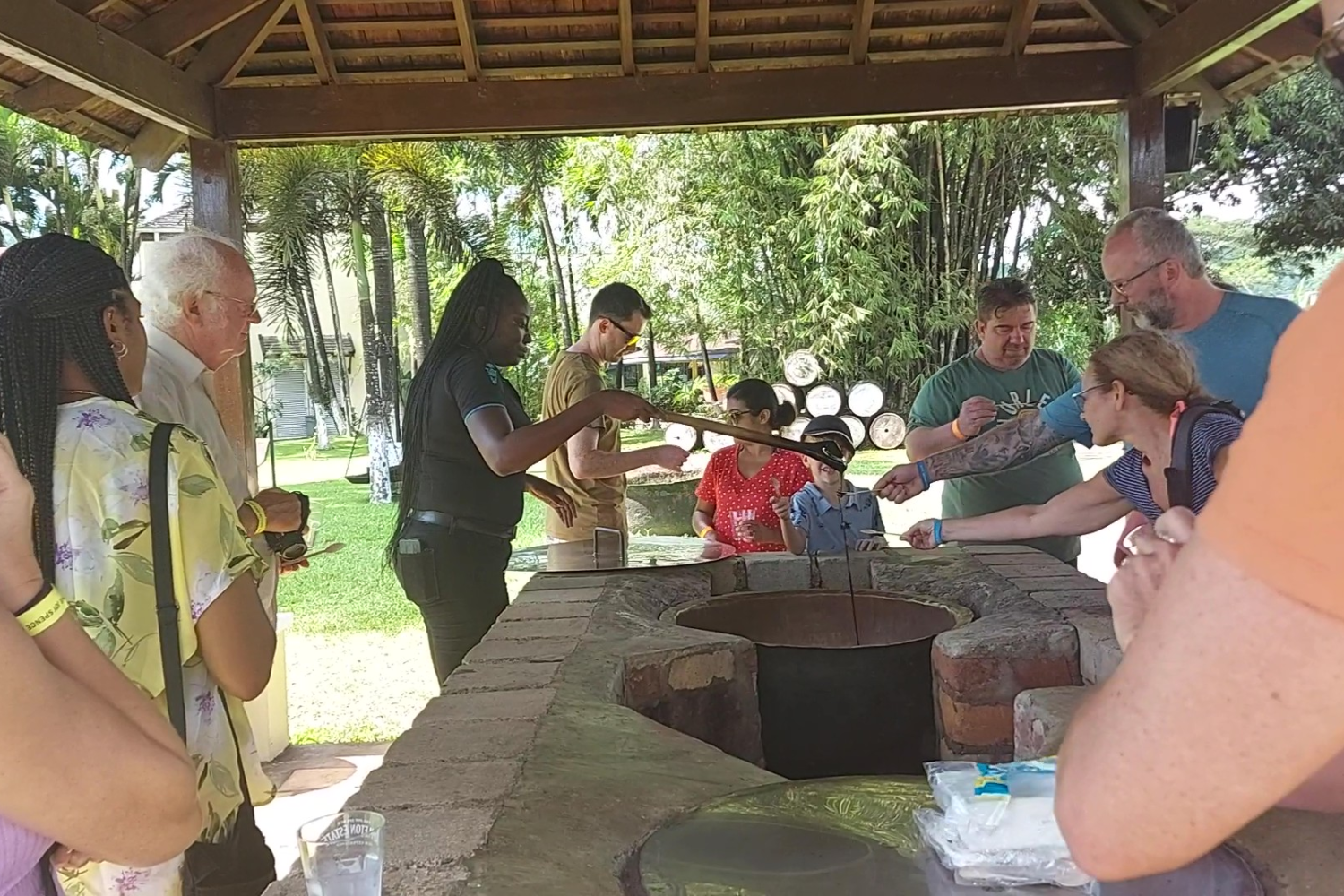Visitors stand around a brick counter as a tour guide dips a wooden spoon into a container of molasses and holds it up, while several guests stretch out their own spoons to collect the dripping molasses for tasting