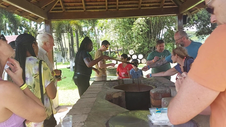 Visitors stand around a brick counter as a tour guide dips a wooden spoon into a container of molasses and holds it up, while several guests stretch out their own spoons to collect the dripping molasses for tasting