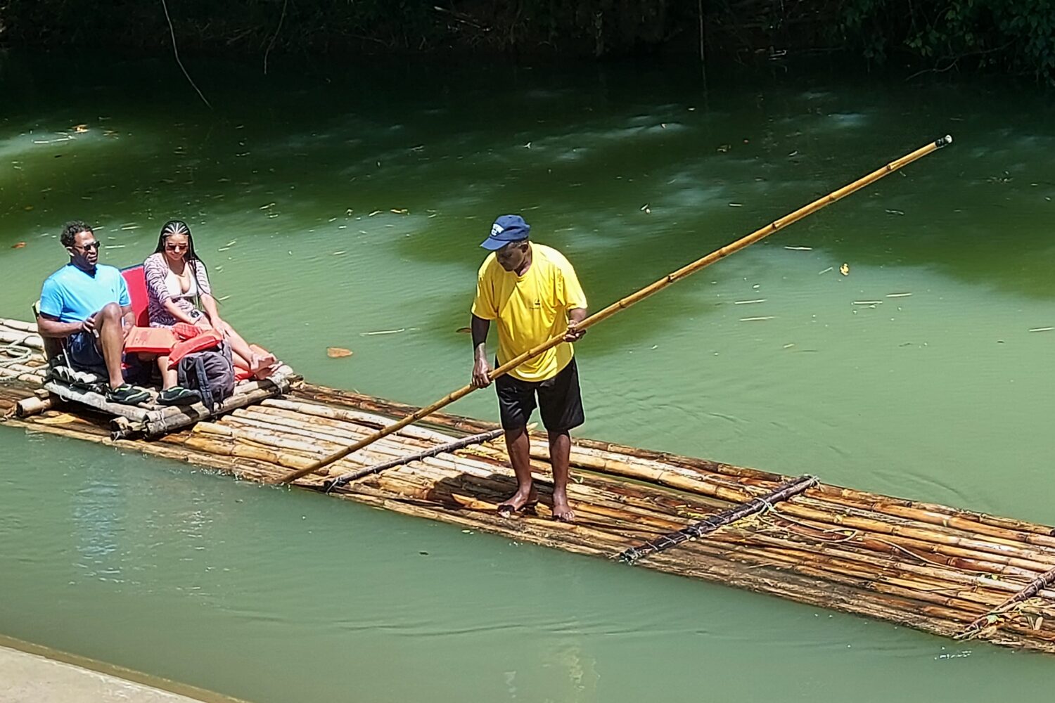 2 people seated and 1 man standing on a bamboo raft on a river