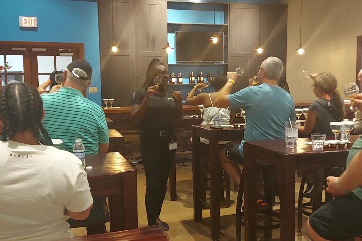 Visitors sit around tables in the Appleton Estate tasting room, holding glasses of rum, while the tour guide stands at the front with a glass in hand, explaining how to view, nose, and identify the rum’s properties.