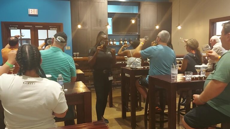 Visitors sit around tables in the Appleton Estate tasting room, holding glasses of rum, while the tour guide stands at the front with a glass in hand, explaining how to view, nose, and identify the rum’s properties.