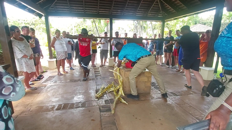 Visitors gather around a manual cane mill, with two guests at each end of the handle turning it in a circle as a staff member feeds sugarcane into the mill, while a tour guide stands nearby giving instructions.
