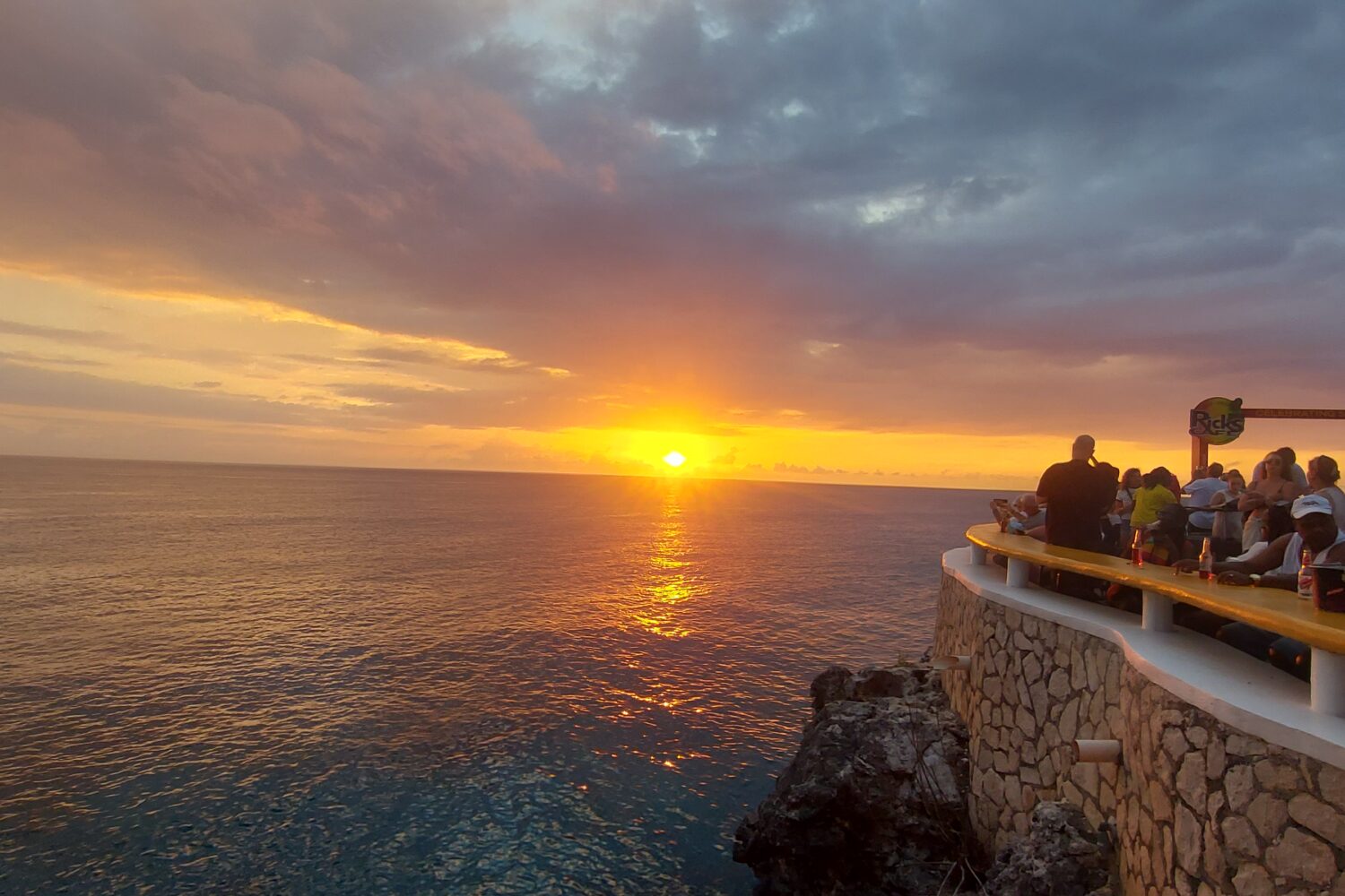 Visitors gather at Rick’s Café in Negril, Jamaica, enjoying the cliffside view as the Caribbean sun sets in vibrant colors, creating an unforgettable travel moment.