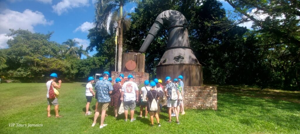 Group of visitors wearing hard hats gathered around a tour guide at Hampden Estate in Trelawny, listening to information about historic pot stills used in rum production.
