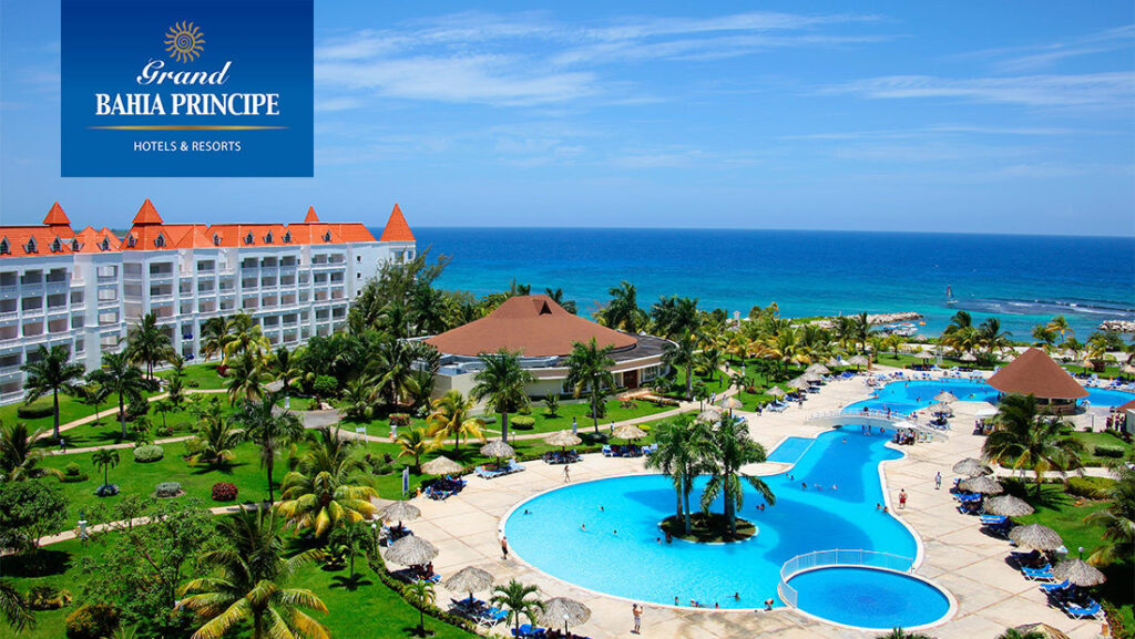Panoramic view of the Grand Bahia Principe Resort in Jamaica with elegant white buildings, lush palm trees, turquoise swimming pools, and the sparkling Caribbean Sea beyond, creating a serene tropical atmosphere.