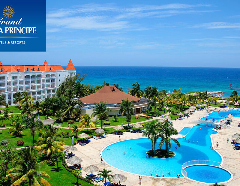 Panoramic view of the Grand Bahia Principe Resort in Jamaica with elegant white buildings, lush palm trees, turquoise swimming pools, and the sparkling Caribbean Sea beyond, creating a serene tropical atmosphere.